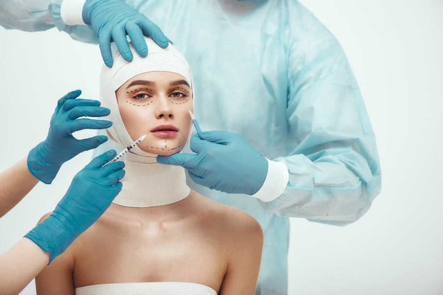 Risks Eyebrow Lift Botox Stop aging. Young attractive woman with head in bandages looking at camera while doctors making plastic surgery operation. Plastic surgeons in blue gloves holding scalpel and syringe near patient face. Facelifting. Cosmetic procedures